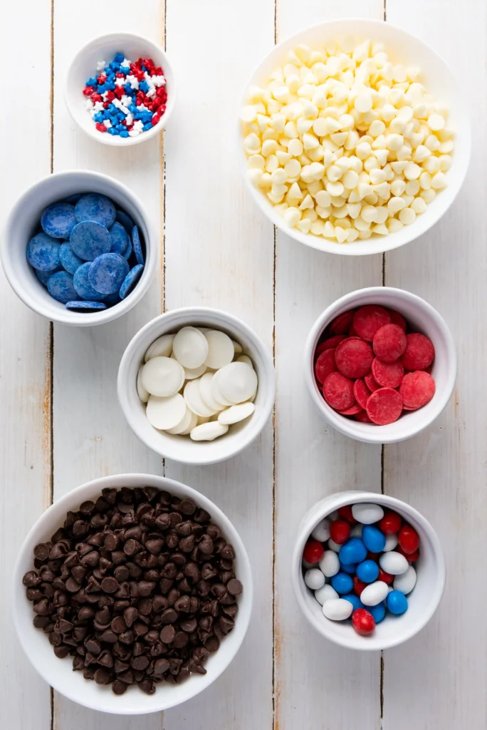 ingredients for the fourth of july dessert - Top-down shot of candy bowls with blue discs, white chocolate chips, red/white/blue sprinkles, red candies, and chocolate chips on a white wooden surface.