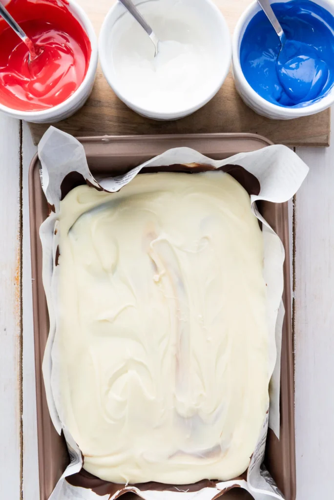 White chocolate in a parchment-lined pan with white frosting, surrounded by red, white, and blue icing bowls.",