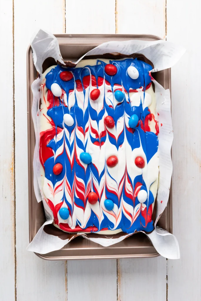 Top-down view of a rectangular pan containing chocolate bark with red, white, and blue marbled icing and scattered red, white, and blue round candies, with parchment paper lining the pan
