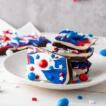 a red, white and blue patriotic dessert -chocolate bark with patriotic m&ms on a white plate