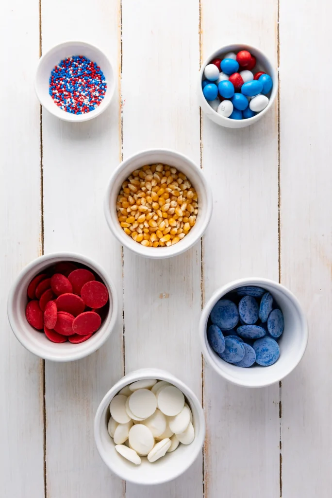 Patriotic Popcorn - Cultivating Brilliant Minds Top-down view of white bowls on a white wooden surface, each filled with different colorful candies (red, white, blue).