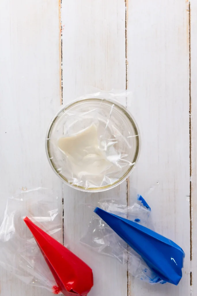 Patriotic Popcorn - Cultivating Brilliant Minds Top-down view of a glass bowl with plastic wrap over pale cream on a white wooden table; red and blue piping bags nearby.
