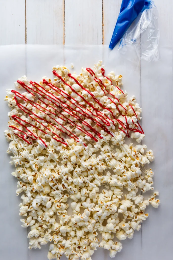 Patriotic Popcorn - Cultivating Brilliant Minds Popcorn scattered on a white wooden surface with red candy drizzle and a blue piping bag in the top-right corner.