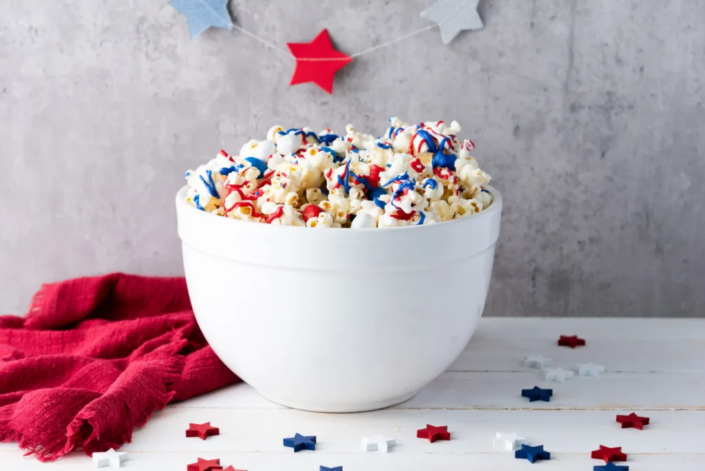 Patriotic Popcorn - Cultivating Brilliant Minds White bowl of popcorn with red, white, and blue sprinkles on a white table, with a red cloth nearby for a patriotic celebration.