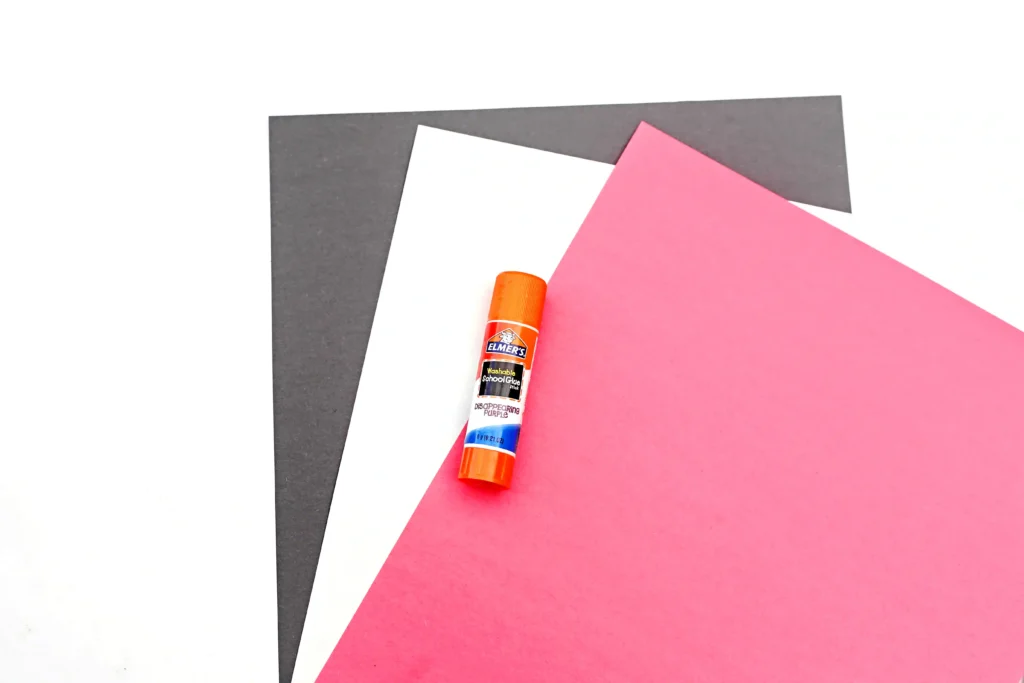 Layered pink, white, and dark gray construction paper with an orange glue stick on top.