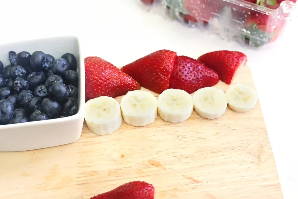 Blueberries in a white bowl beside sliced strawberries and banana rounds on a wooden surface.