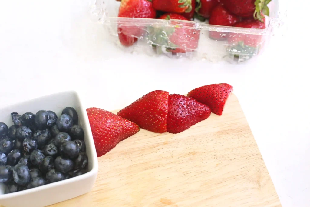 Close-up of blueberries in a white bowl with sliced strawberries on a wooden cutting board.