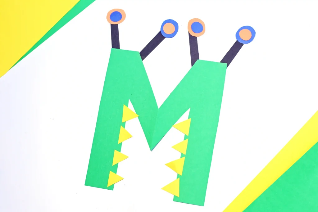 a letter m craft made out of construction paper with a green letter m that has eyes and teeth to look like a monster