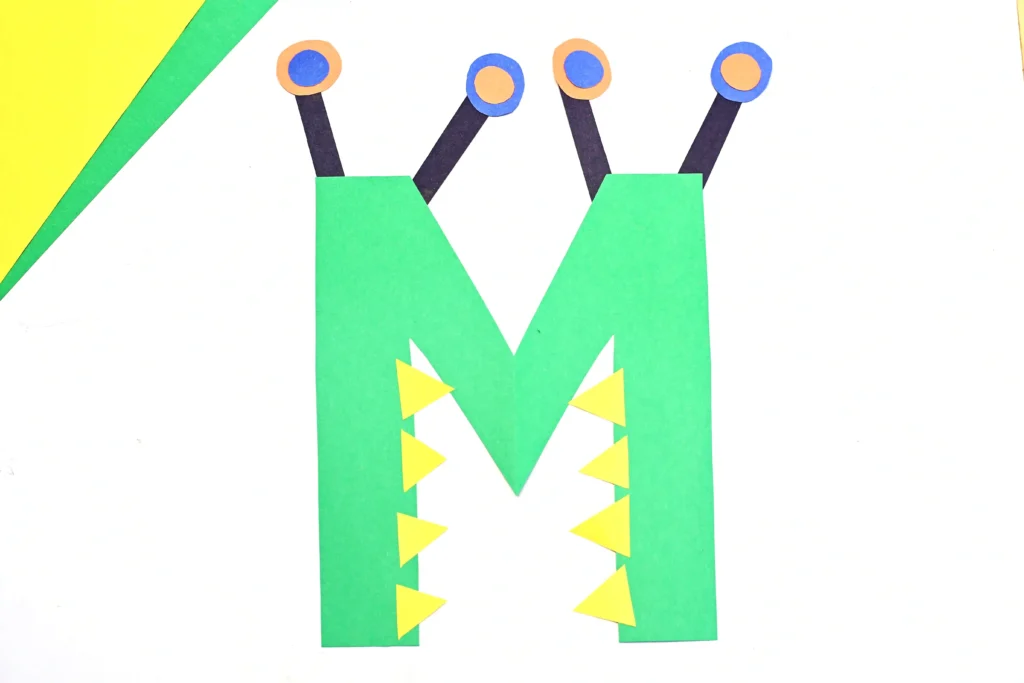 a letter m monster craft made out of construction paper with a green letter m that has eyes and teeth