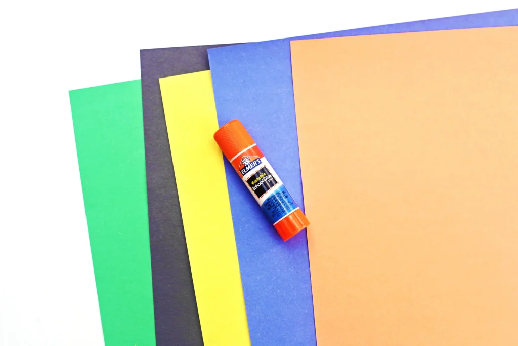 supplies for this alphabet craft: Assorted colored construction paper sheets overlapped with a orange glue stick resting diagonally across them (green, yellow, blue, orange).