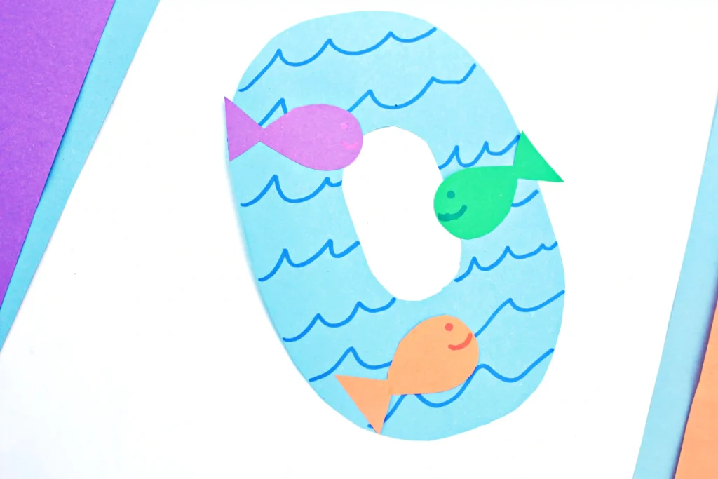 a letter O cut out of blue paper with blue waves drawn on and colorful paper fish glued on top