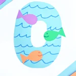 a letter O cut out of blue paper with blue waves drawn on and colorful paper fish glued on top to make a letter activity for kids