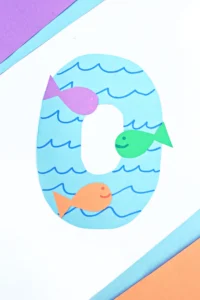 a letter O cut out of blue paper with blue waves drawn on and colorful paper fish glued on top to make a letter activity for kids