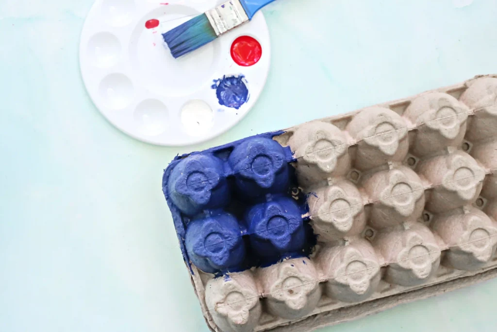 Paint palette with blue and red paint and a brush, next to a blue-painted egg carton.