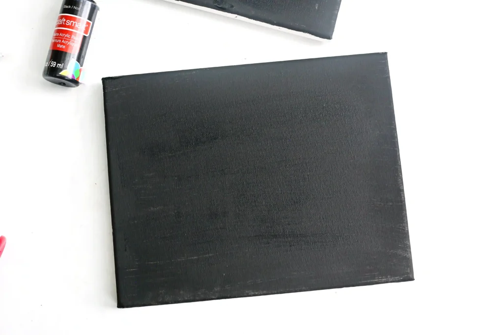 Partial view of black canvases with a CraftSmart acrylic paint bottle on a white surface.