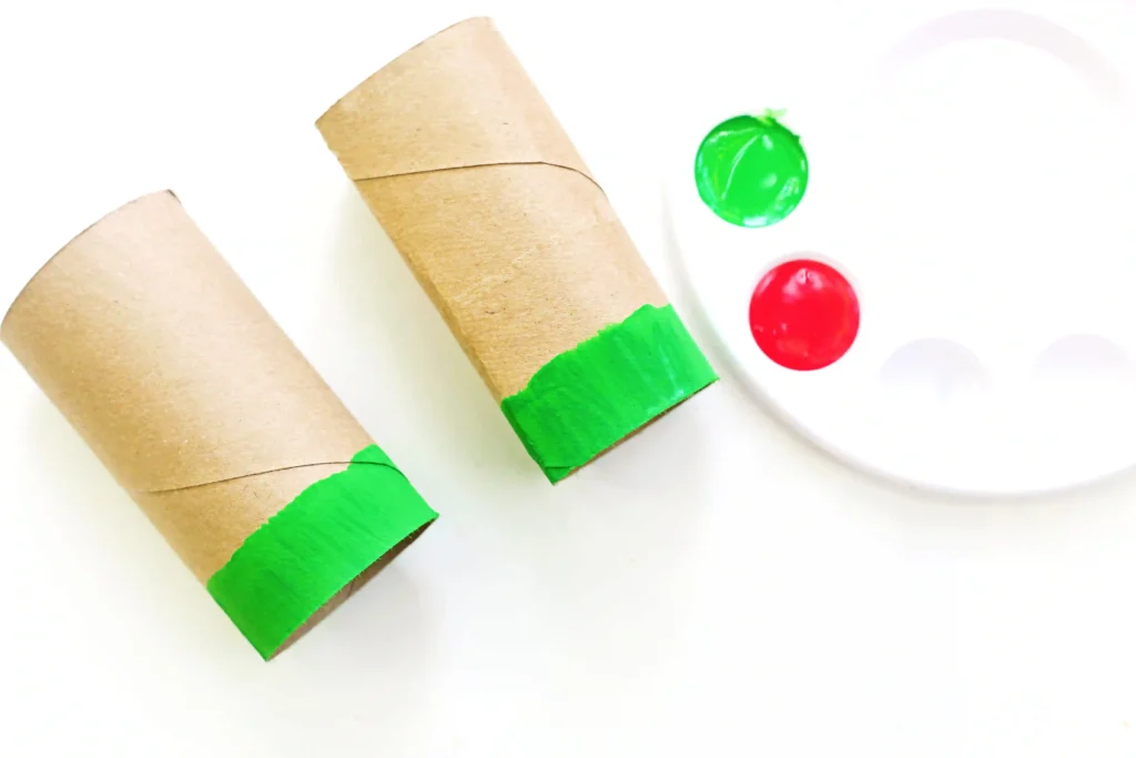 Two brown cardboard tubes painted green sit on a white surface beside green and red paint spots, suggesting a craft project.