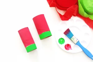 Colorful art supplies on a white surface: red toilet paper tubes with green ends, a blue paintbrush, a green paint lid, and a white palette with red paint.