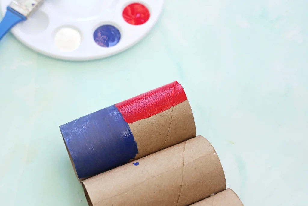 Toilet Paper Roll American Flag - Cultivating Brilliant Minds Close-up of painted cardboard tubes in blue and red, with a white paint palette showing blue and red wells in the blurred background.