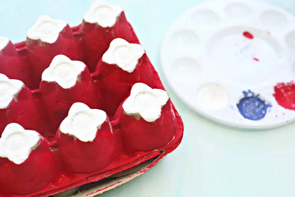 Close-up of a red and white painted cardboard egg carton with white scalloped edges against a light blue background.
