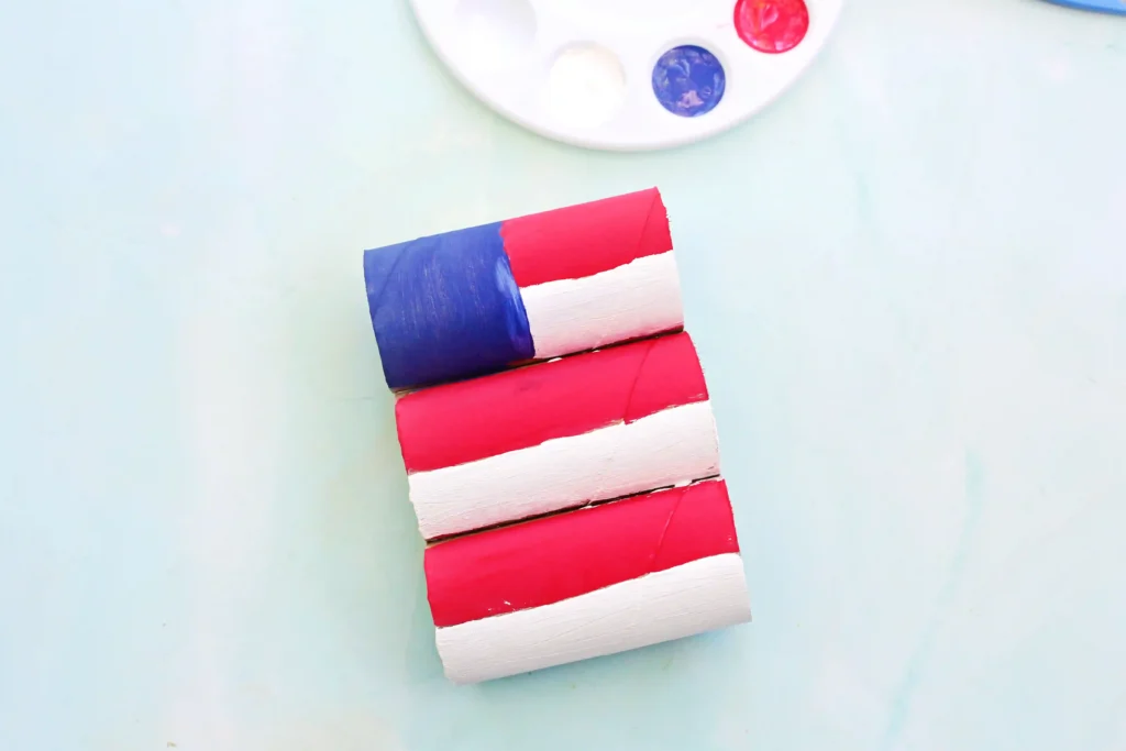 Toilet Paper Roll American Flag - Cultivating Brilliant Minds Stack of small painted toilet paper rolls in red, white, and blue leaning diagonally on a pale blue background with a partial white paint palette overhead.