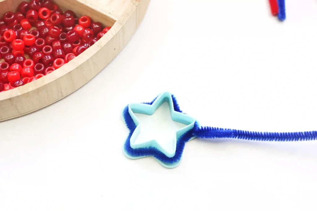 Close-up of red plastic beads in a wooden circular tray, with a blue star-shaped cutter and blue pipe cleaners on a white surface.
