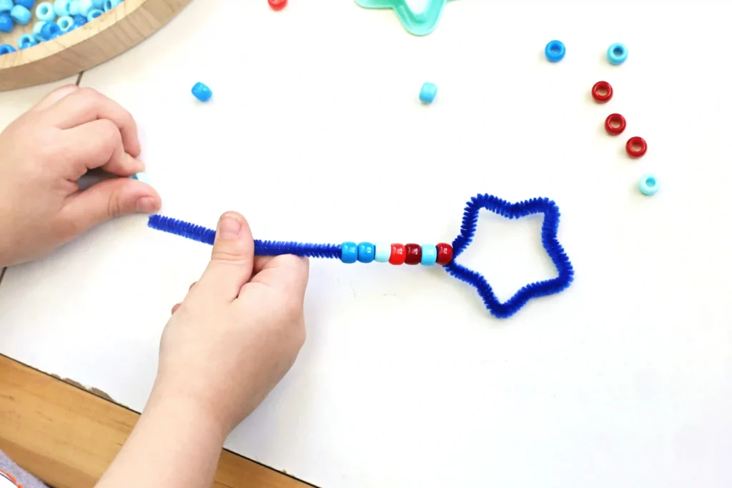 Child's hands thread light blue beads onto a blue pipe cleaner during a craft project with a nearby tray of blue beads.