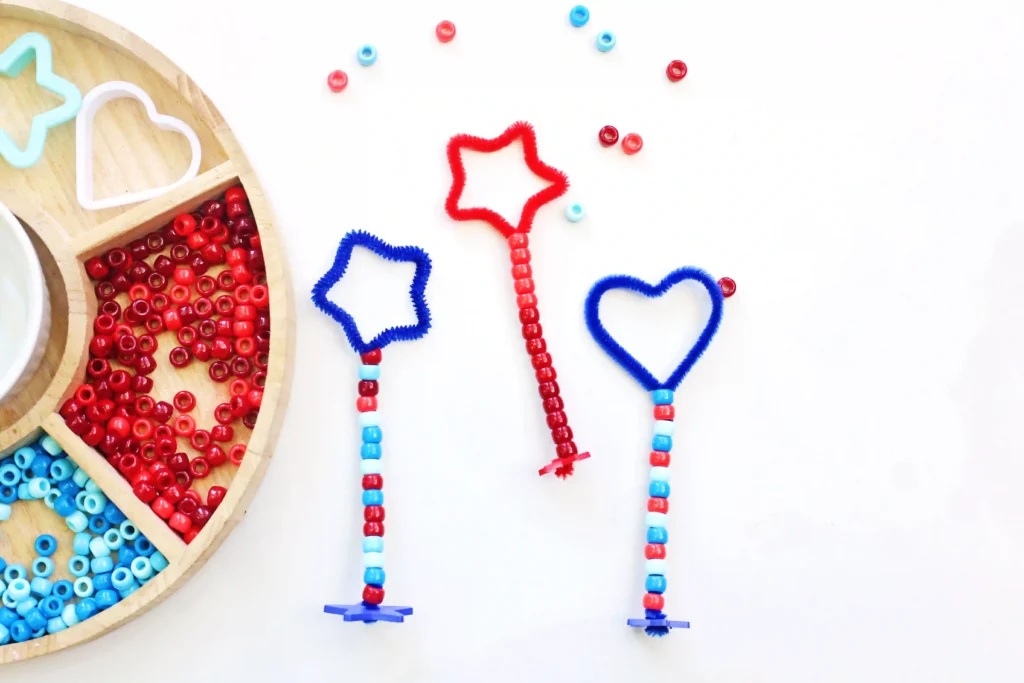 patriotic bubble wands: red and blue star and heart shapes with bead wands on a white surface beside a wooden bead tray teeming with small red beads.