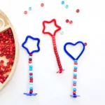 patriotic bubble wands: red and blue star and heart shapes with bead wands on a white surface beside a wooden bead tray teeming with small red beads.