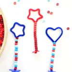 Beaded wands with red and blue pipe-cleaner shapes (star and heart) hanging from striped bead strings, with a wooden tray of beads visible on the left.
