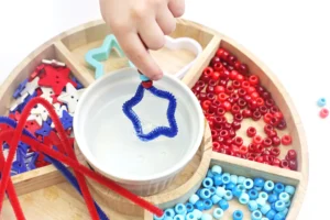 Hand dips a blue pipe cleaner into a cup of bubble solution, with red and blue beads and star-shaped pieces in a wooden tray nearby.