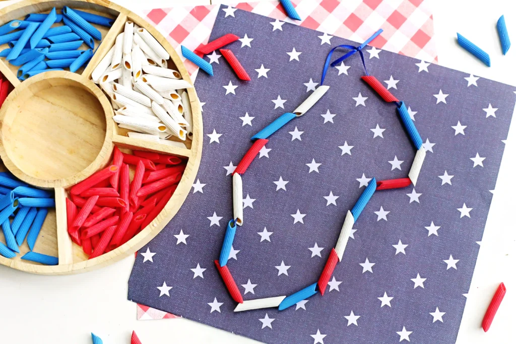 Patriotic Pasta Necklaces - Cultivating Brilliant Minds Craft supplies arranged on a star-patterned surface: red, white, and blue pasta pieces are strung together to create a necklace