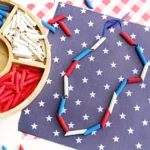 Patriotic craft scene with red, white, and blue penne arranged on starry blue paper; paper bead necklace adds a festive touch.