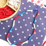 Patriotic craft scene with red, white, and blue penne arranged on starry blue paper; paper bead necklace adds a festive touch.