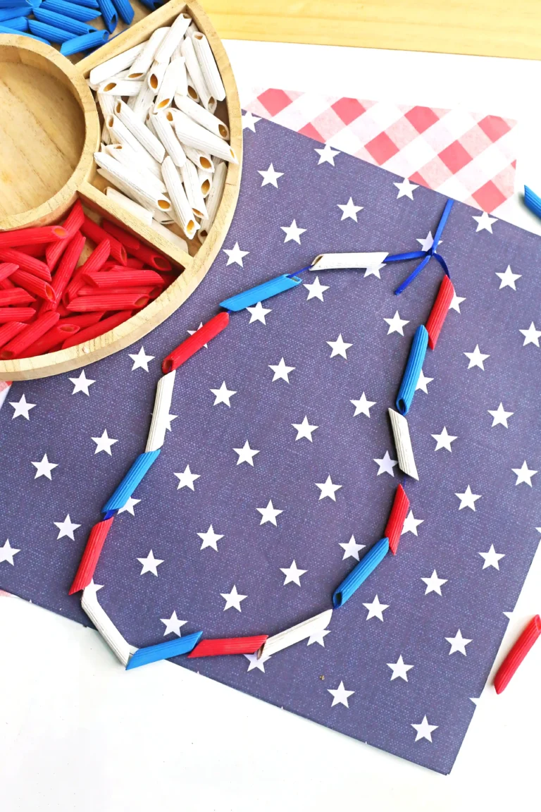 Patriotic craft scene with red, white, and blue penne arranged on starry blue paper; paper bead necklace adds a festive touch.