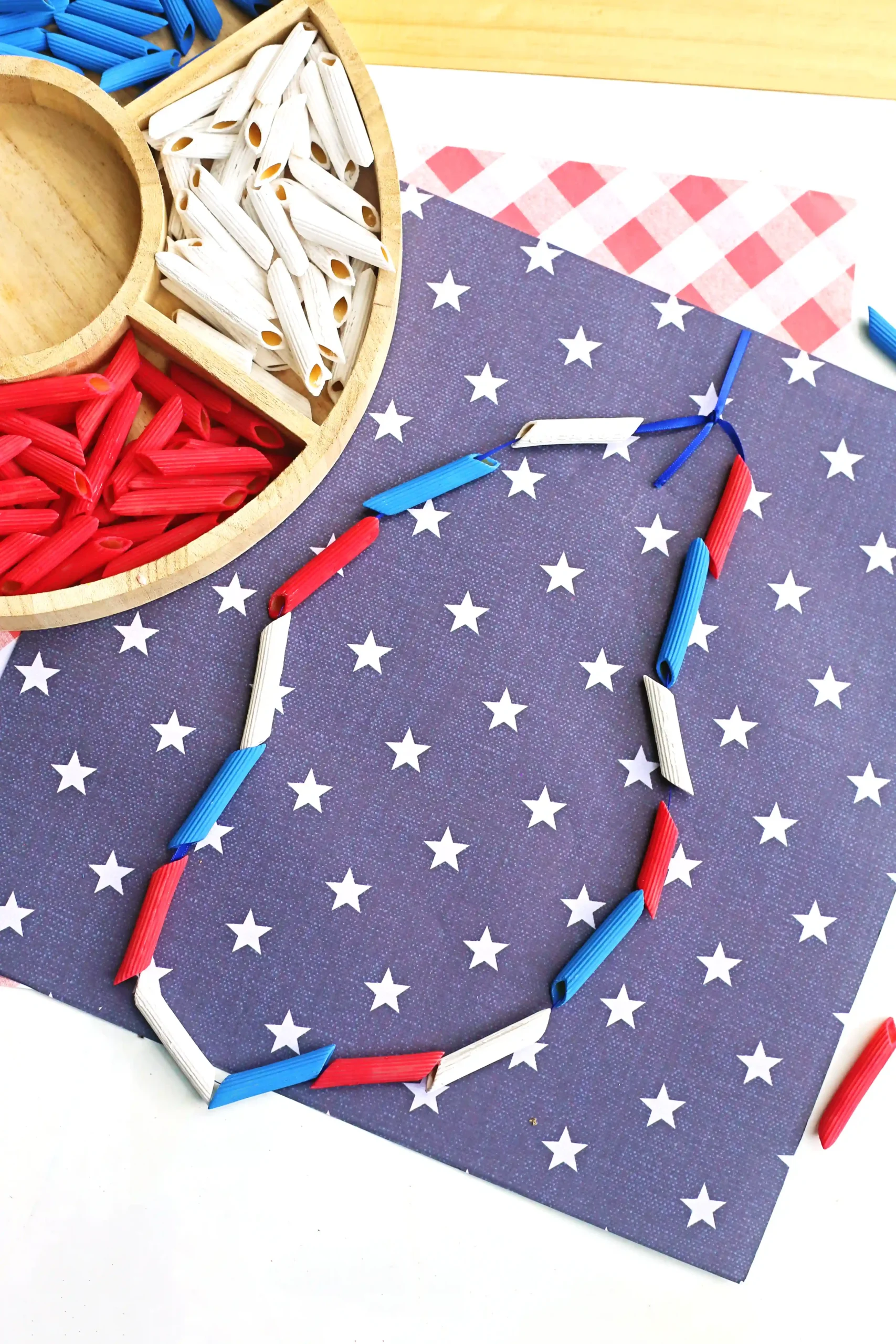 Patriotic craft scene with red, white, and blue penne arranged on starry blue paper; paper bead necklace adds a festive touch.