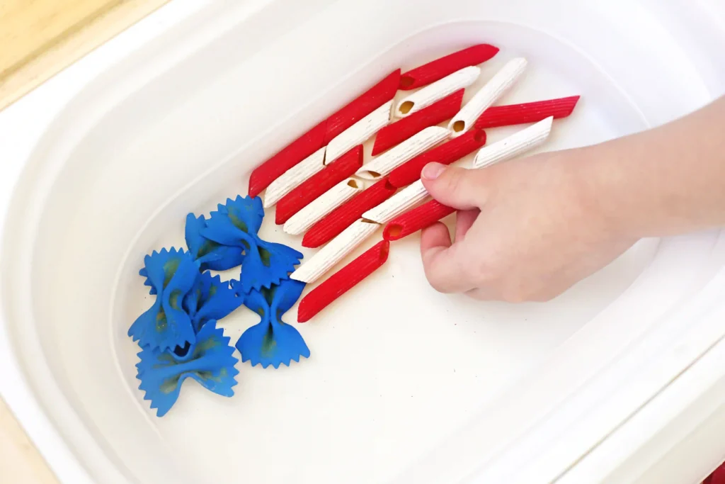Hand arranging red and white dyed pasta on a white plate, with blue decorative pieces nearby for a craft project.