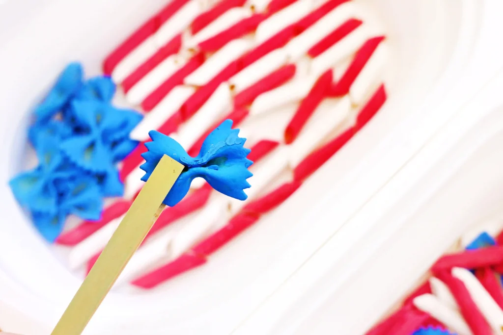 tongs holding farfalle pasta that is dyed blue with a patriotic pasta sensory bin in the background