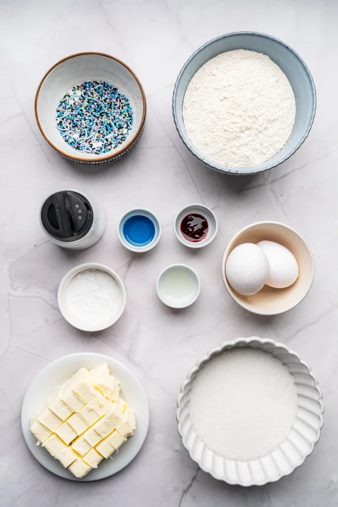 Overhead shot of baking prep: large bowl of flour, sprinkle-topped bowl, eggs, butter, sugar, and small bowls on a marble surface.