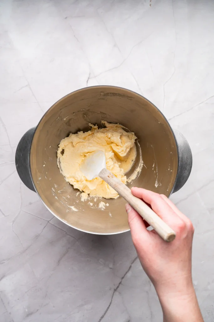 Top-down view of a stainless steel mixing bowl with butter being stirred by a wooden spoon, held by a hand over a marble countertop.
