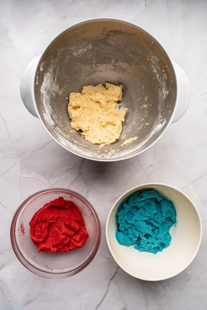 Top-down view of a metal mixing bowl with pale dough, surrounded by bowls of red and turquoise dough on a light marble counter