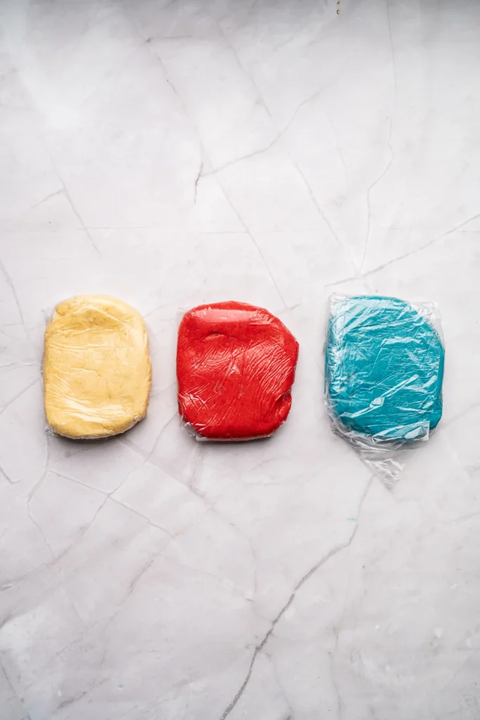 Three colorful wrapped rectangular dough in yellow, red, and blue lined up on a light marble surface.