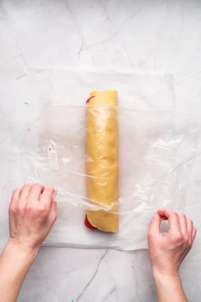 Hands wrap parchment around a long dough log on a marble surface, with a light wooden rolling pin nearby.