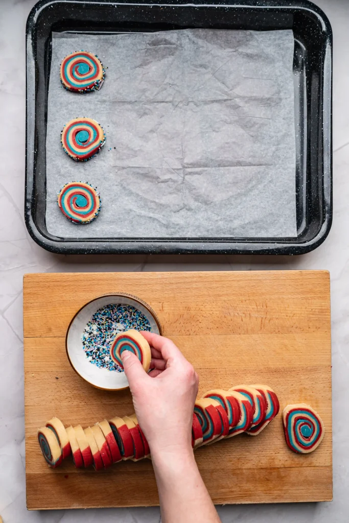 A hand dips a rainbow spiral cookie into a bowl of multicolored sprinkles on a wooden cutting board to complete the patriotic cookie recipe
