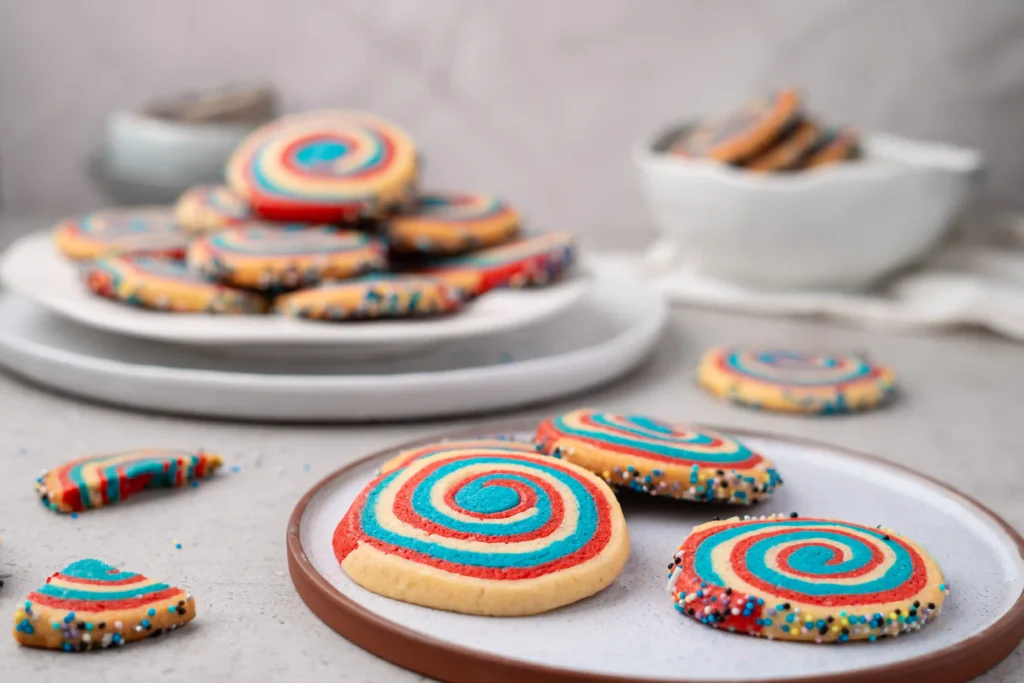 Plate of rainbow swirl cookies with blue, red, and yellow icing on a serving platter, with more cookies scattered on a light table in the blurred background.
