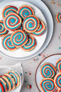 red white and blue swirled patriotic pinwheel cookies with sprinkles on the sides on a white plate