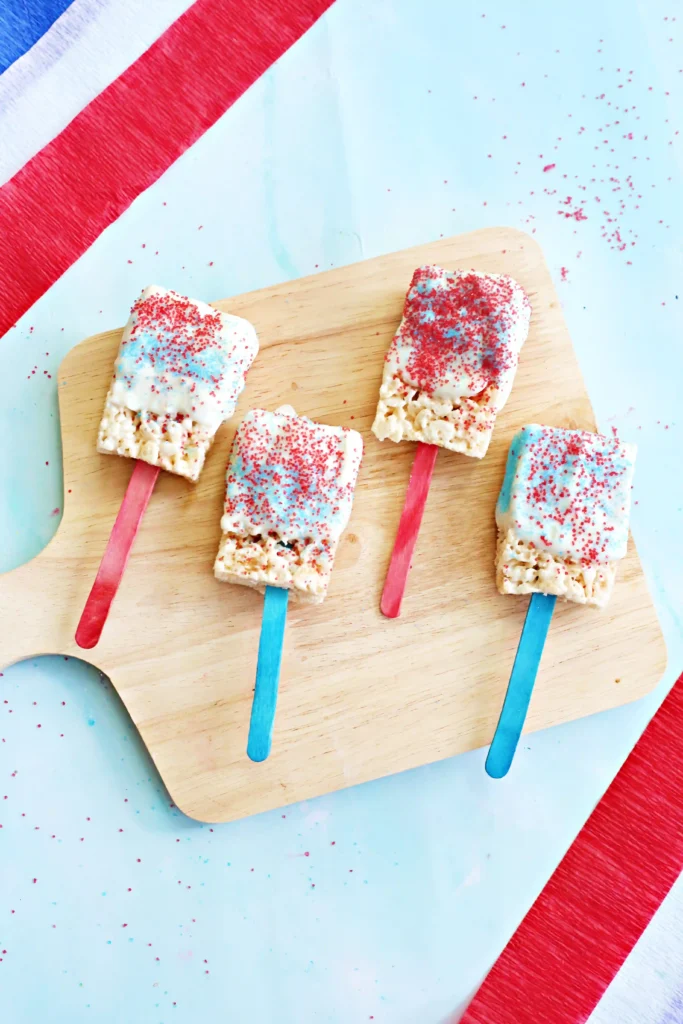 Patriotic Rice Krispie Pops - Cultivating Brilliant Minds patriotic rice krispie treats on red and blue stick with melted white chocolate on top and red and blue sprinkles