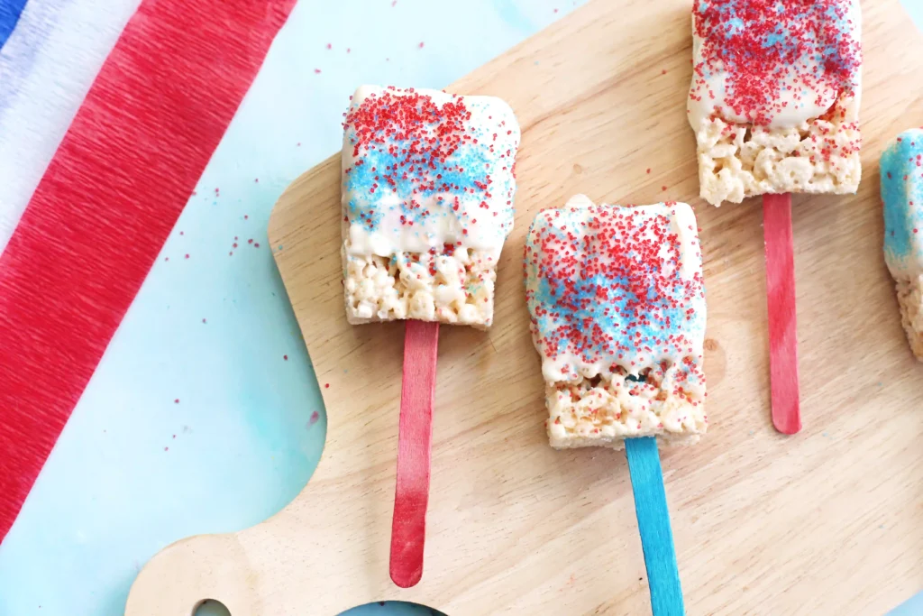 Patriotic Rice Krispie Pops - Cultivating Brilliant Minds Patriotic rice cereal treats on red and blue sticks with white icing and red sprinkles, arranged on a wooden board.