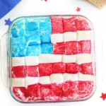 Patriotic marshmallow squares in red, white, and blue arranged in a glass dish with star confetti and crackers to the side.