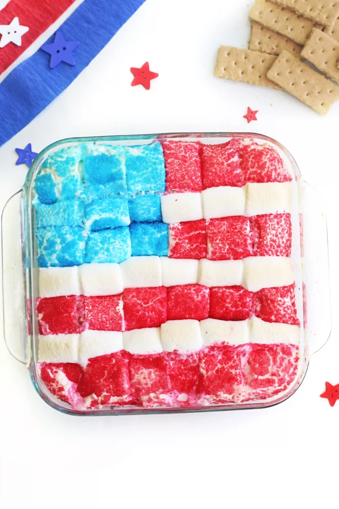 Patriotic marshmallow squares in red, white, and blue arranged in a glass dish with star confetti and crackers to the side.