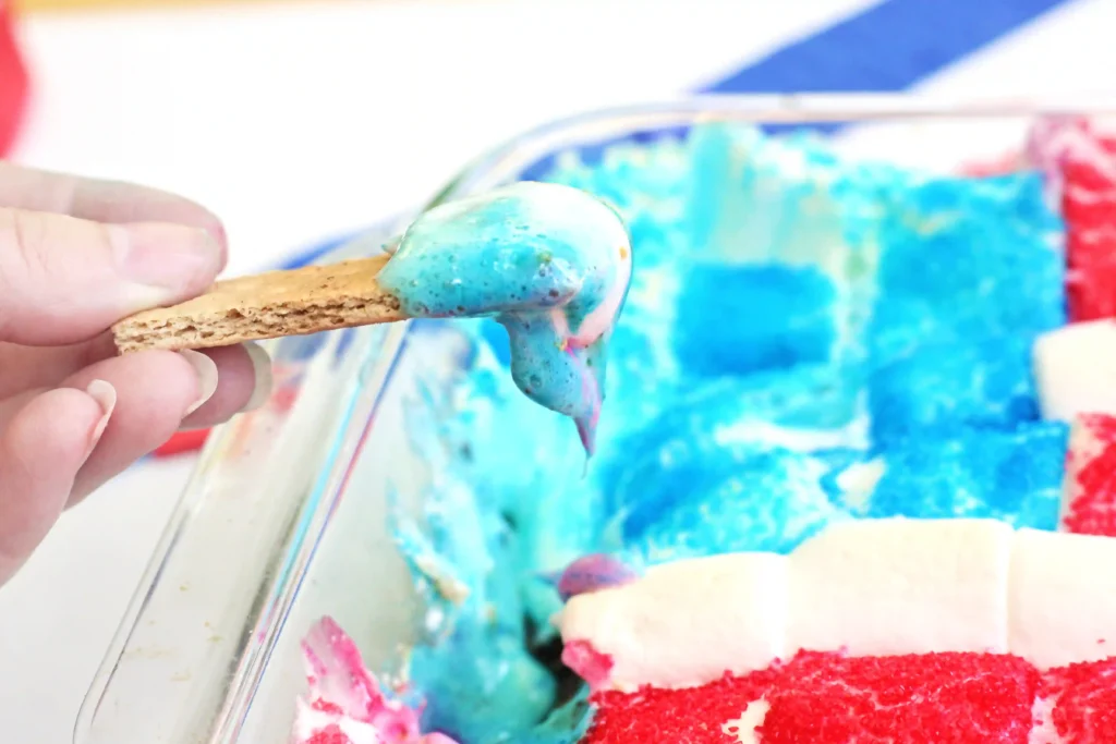 Close-up of a finger holding a graham cracker dipped in marshmallows with blue sanding sugar
